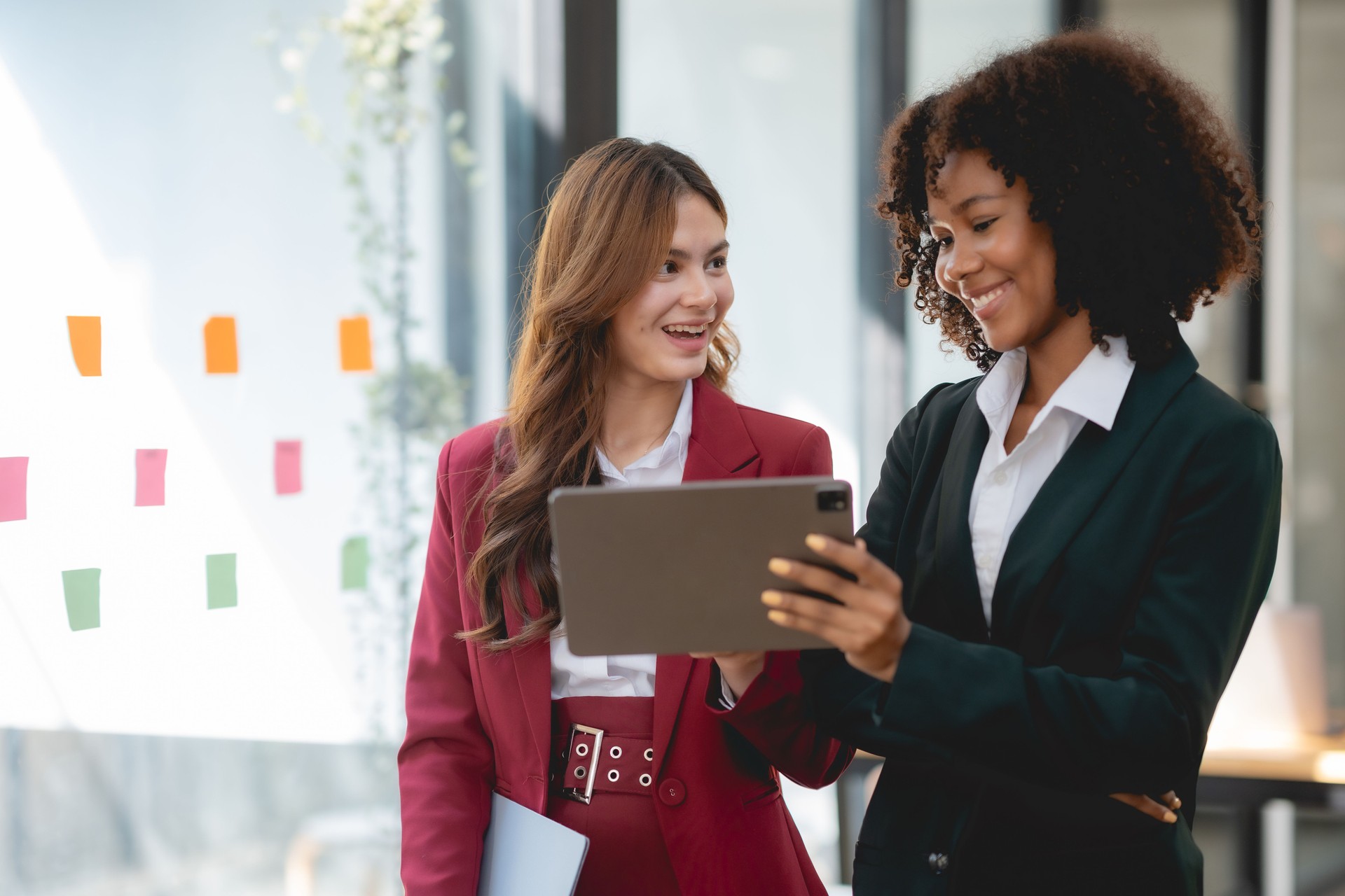 Two young business woman using a digital tablet while standing in a boardroom. Two happy businesswoman talking and consulting working together in the office.